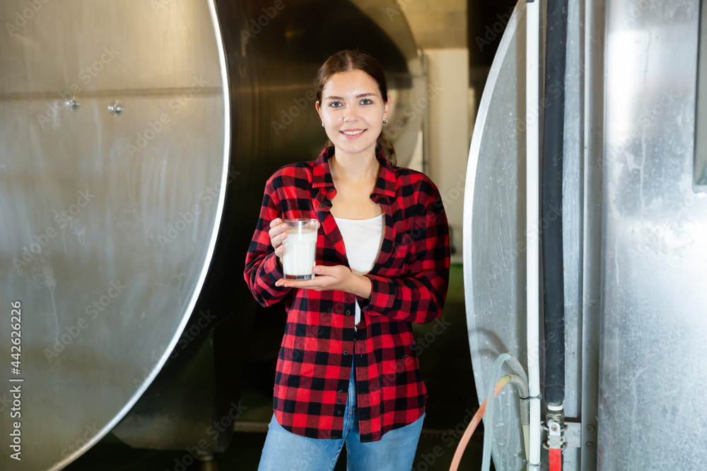 Smiling young workwoman standing near large tanks in storage room of dairy farm, holding glass with fresh milk