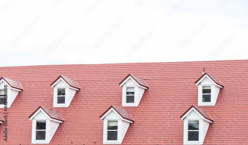 The corner of a red metal shingled roof with two raised dormers and three sunken small single windows in the exterior wall of a white stucco building. The vintage building has a clean design.