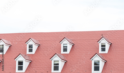 The corner of a red metal shingled roof with two raised dormers and three sunken small single windows in the exterior wall of a white stucco building. The vintage building has a clean design.