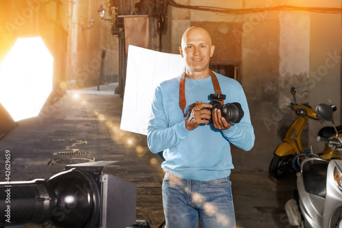 Portrait of male photographer standing with camera among professional photo equipment on old city street