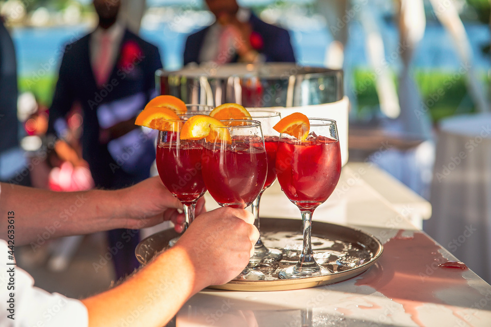 Hands holding red cocktails with orange slices. Party or wedding reception with alcohol drinks, cocktail hour. Blurred background