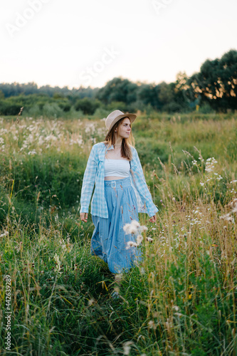 Beautiful redhead girl in standing in a centre of a green summer meadow, wearing a straw hat and long skirt