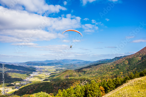 紅葉の山に囲まれた中で青空に向かって飛ぶパラグライダー。京都府与謝郡与謝野町