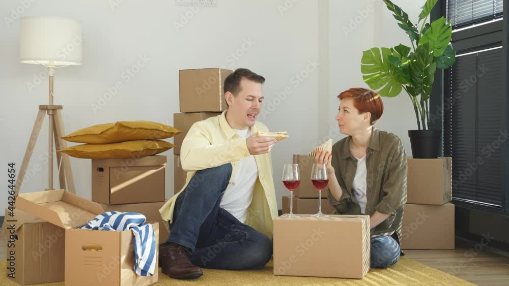 Cheerful Couple Eat Sandwich, Drinking Red Wine Celebrating Relocation And Moving Into New House While Sitting On Floor With Unpacked Cardboard Boxes, Romantic Time, Excited by Purchase Cheerful Couple Eat Sandwich, Drinking Red Wine Celebrating Relocation And Moving Into New House While Sitting On Floor With Unpacked Cardboard Boxes, Romantic Time, Excited by Purchase
