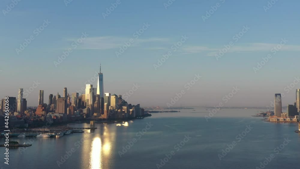 An aerial view of lower Manhattan and New Jersey from over the Hudson River at sunrise. The sun reflects on a few buildings while the camera dolly out over the calm river and a red barge is revealed.