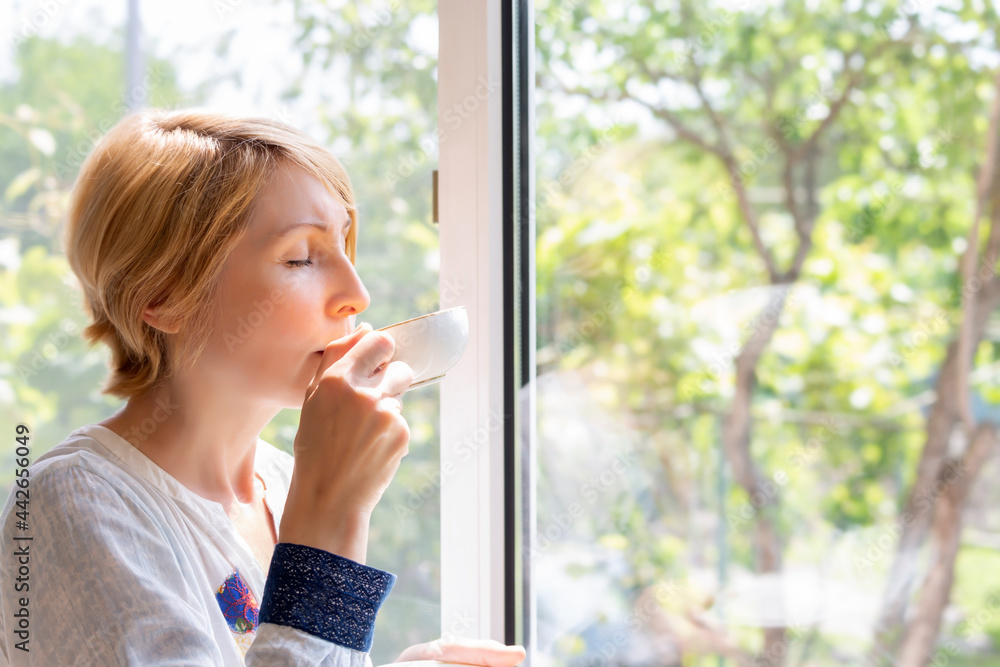 Fototapeta premium A woman drinking tea at the picture window with her eyes closed with pleasure