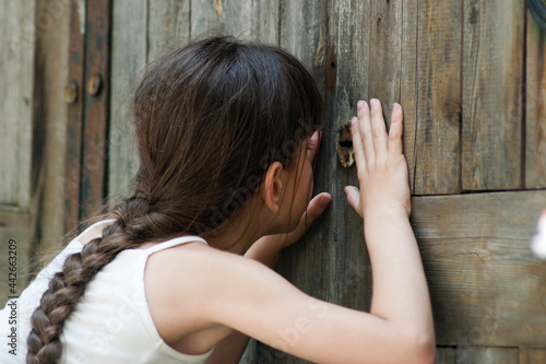The child peeps through a keyhole. The little girl (eight years old) looks through a keyhole of old wooden gate. Outdoors