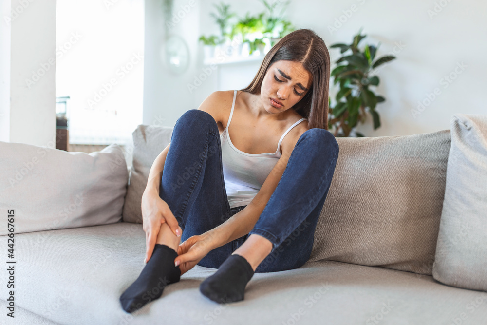 Hands massaging swollen foot while sitting on sofa during the day at home. Photo of Young Caucasian woman suffering from pain in leg. Woman massaging her legs after all day at work in office Hands massaging swollen foot while sitting on sofa during the day at home. Photo of Young Caucasian woman suffering from pain in leg. Woman massaging her legs after all day at work in office