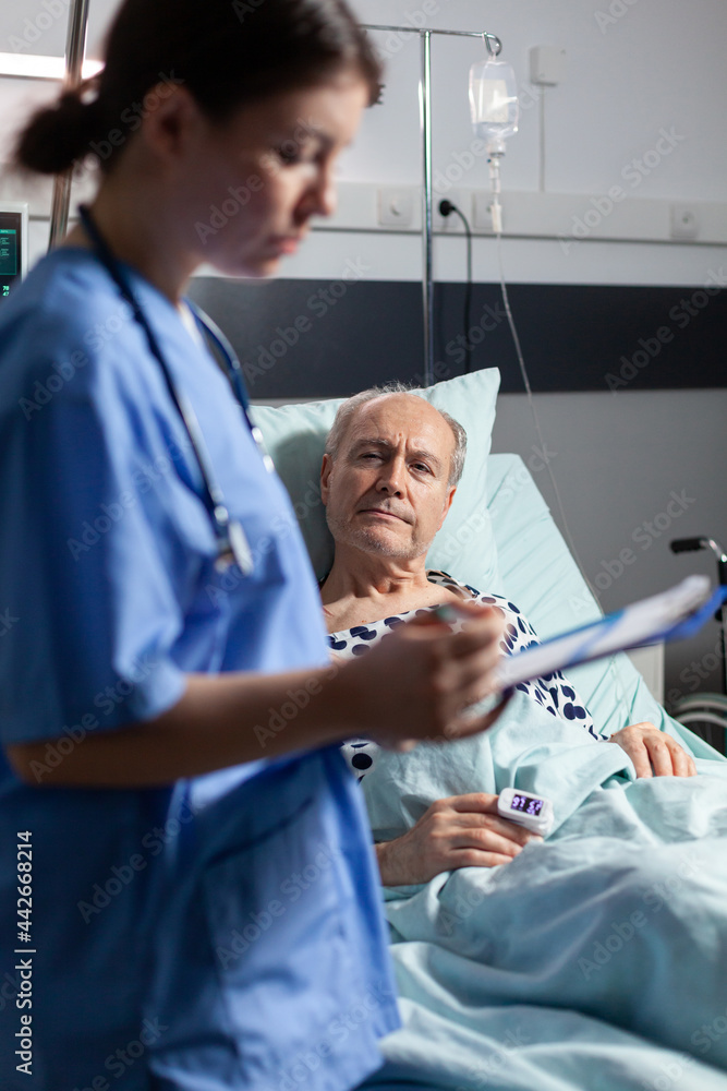 Medical nurse in scrubs taking notest on cliboard after consultation of sick unwell senior man. Patient with iv drip attached breathing with help from oxygen mask. Medical nurse in scrubs taking notest on cliboard after consultation of sick unwell senior man. Patient with iv drip attached breathing with help from oxygen mask.
