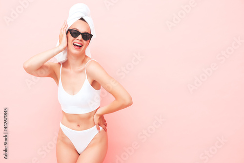 Young beautiful smiling woman in white lingerie. Sexy carefree model in underwear and towel on head posing pink wall in studio. Positive and happy female enjoying morning in sunglasses