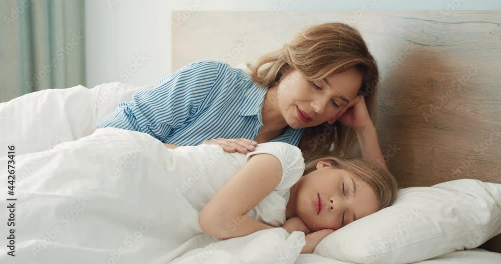 Happy Caucasian mother lies next to her sleeping daughter in bed and tries to gently wake her in the morning. Happy Caucasian mother lies next to her sleeping daughter in bed and tries to gently wake her in the morning.