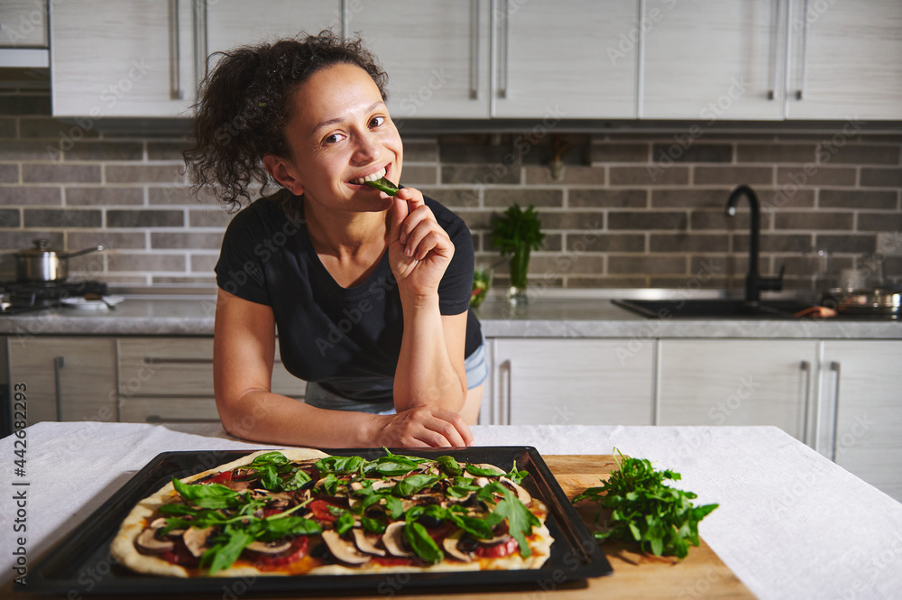 Smiling Latin-American woman leaning on kitchen table with hot pizza and tasting basil, smiling, posing at camera over home kitchen background