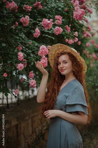 Beautiful redhead freckled woman with long curly natural hair, wearing straw hat, blue dress, posing in blooming rose garden. Beauty, hair care, summer lifestyle concept. 