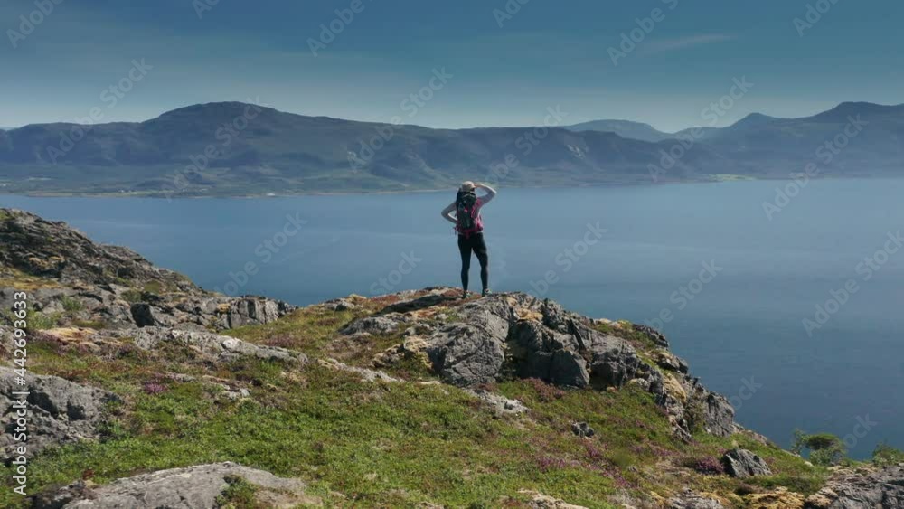 An astounding view opens up in front of a woman hiker who has climbed up the hill: a mountain ridge stretched to the horizon. It is dividing calm fjord water and sky, both of deep blue color. An astounding view opens up in front of a woman hiker who has climbed up the hill: a mountain ridge stretched to the horizon. It is dividing calm fjord water and sky, both of deep blue color.