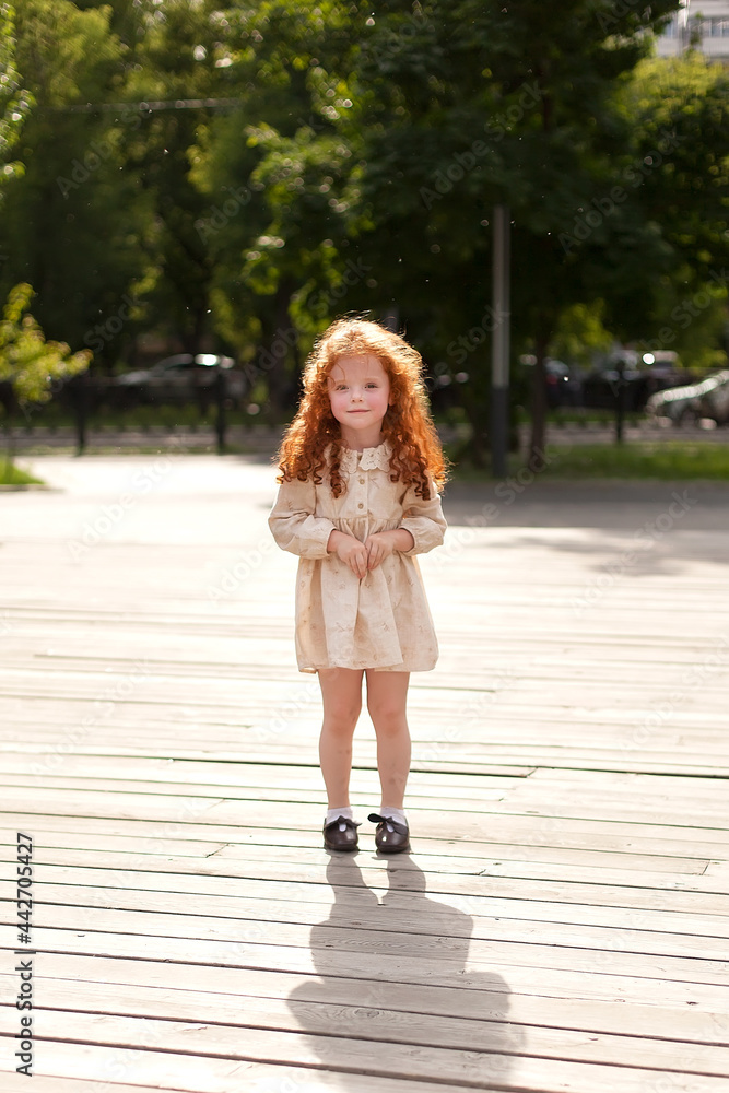 A cute little girl with red hair and a light dress is walking outdoors. Portrait of a small child. Walking in the city park.
