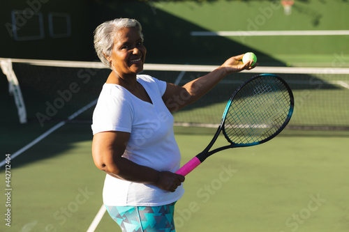 Portrait of smiling senior african american man holding tennis racket and ball on tennis court
