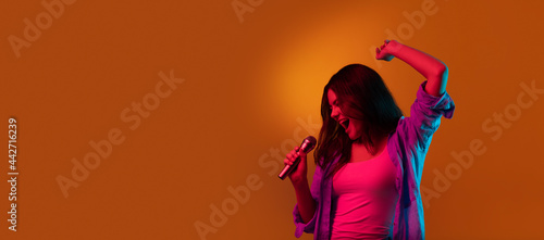 Caucasian female singer portrait isolated on orange studio background in neon light. Beautiful female model in casual style with microphone. Concept of human emotions