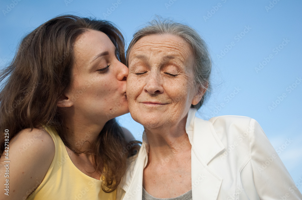 Senior mother with gray hair with her adult daughter looking at the camera in the garden and hugging each other during sunny day outdoors Senior mother with gray hair with her adult daughter looking at the camera in the garden and hugging each other during sunny day outdoors