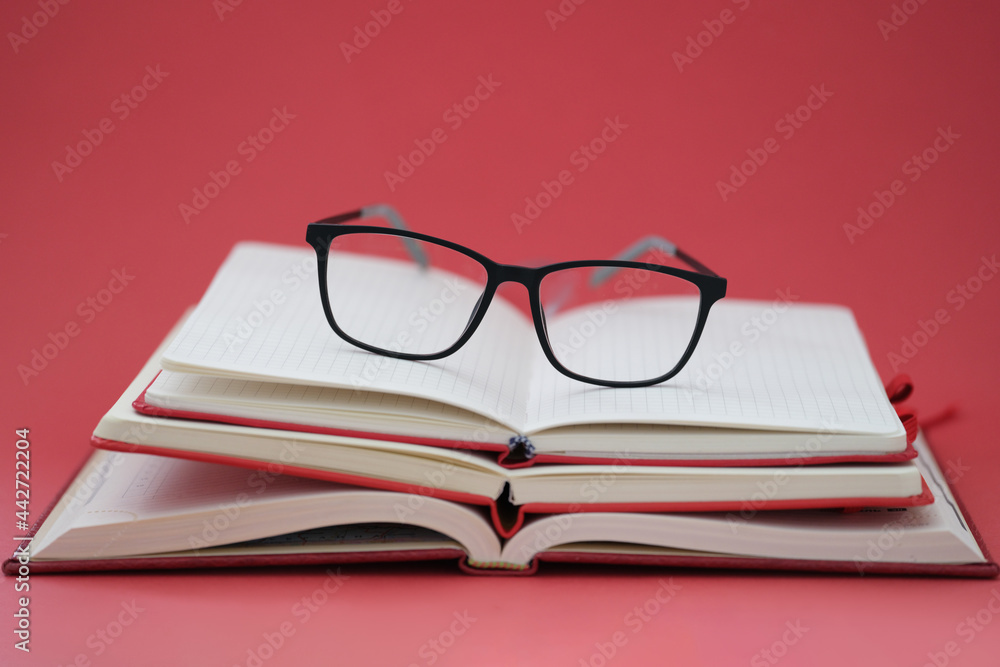 Glasses for sight lying on pile of notebooks on red background closeup Glasses for sight lying on pile of notebooks on red background closeup