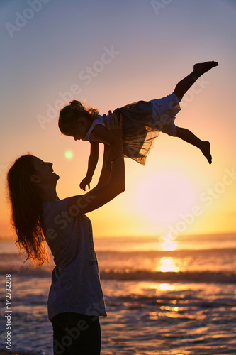 Silhouette of mom and daughter on the beach. Happy Woman throws up a child. A mother plays with her child at dusk and dawn by the ocean. Family travel