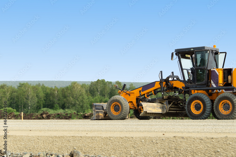 An orange grader is busy with road repairs. Excavator for road works. Road machinery works against the background of the forest. An orange grader is busy with road repairs. Excavator for road works. Road machinery works against the background of the forest.