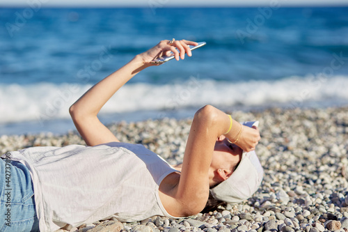  Young woman is lying in a hat with a phone on the beach near the sea is making a selfie. Resting, traveling and relaxing concept. Copy space