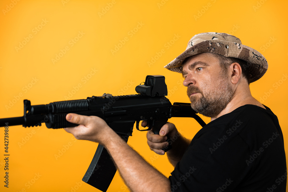 Middle aged man with rifle in black t-shirt and hat against yellow background.