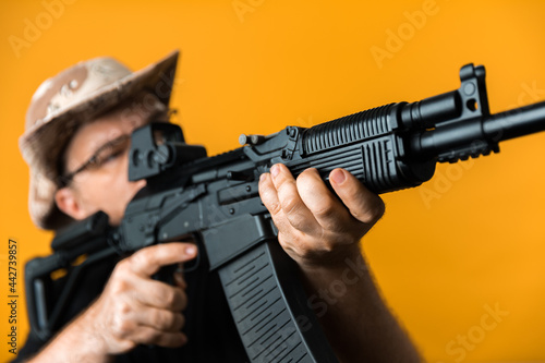 Middle aged man with rifle in black t-shirt and hat against yellow background.