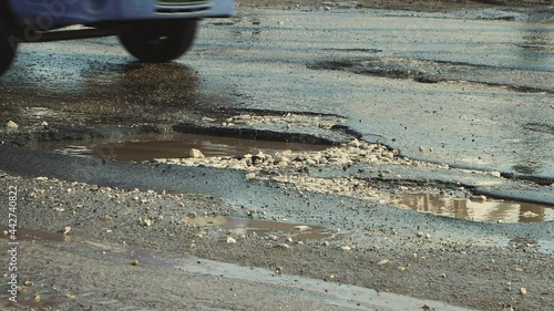 Cars drive in close-up on a bad, bumpy road with holes. Large potholes after rain, damaged road infrastructure. Asphalt in violation of technology. Taxpayer money is wasted and corruption is rampant.