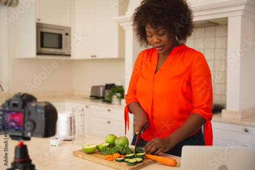 Happy african american woman standing in kitchen preparing food, making vlog using laptop and camera