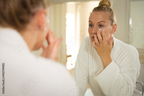 Caucasian woman in bathroom wearing bathrobe, looking in mirror and moisturising face