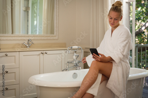 Smiling caucasian woman sitting in bathroom wearing bathrobe holding smartphone, running a bath