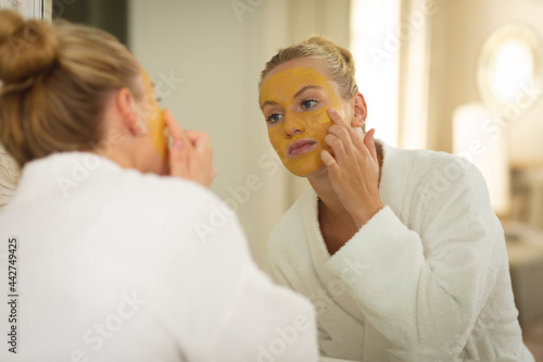 Caucasian woman in bathroom wearing bathrobe, looking in mirror and applying face mask