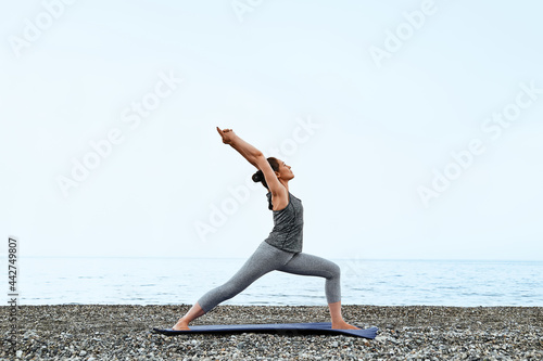 Slim Woman doing Virabhadrasana or warrior pose on the beach in the morning. Strengthening muscles of legs and coordination of body. Practicing yoga outdoors.