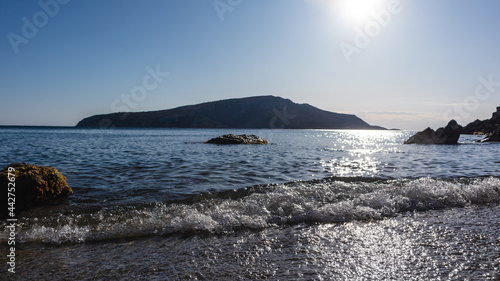 Sun shining on wild shore with calm waves. Mediterranean sea with rocks silhouettes, clear water and sunbeam. Travel Greece near Athens. Summer seaside nature. Blue filter