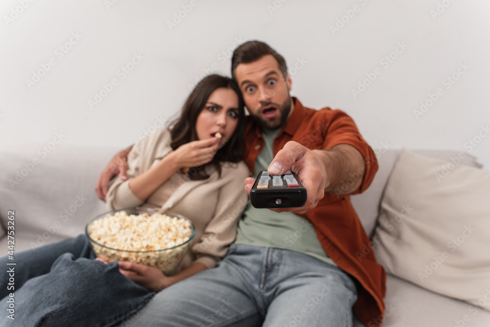 Remote controller in hand of blurred man near amazed girlfriend with popcorn Remote controller in hand of blurred man near amazed girlfriend with popcorn