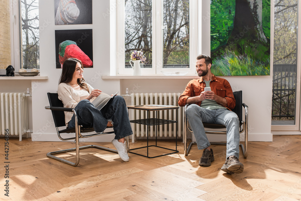 Obraz premium Smiling man with smartphone looking at girlfriend with book at home