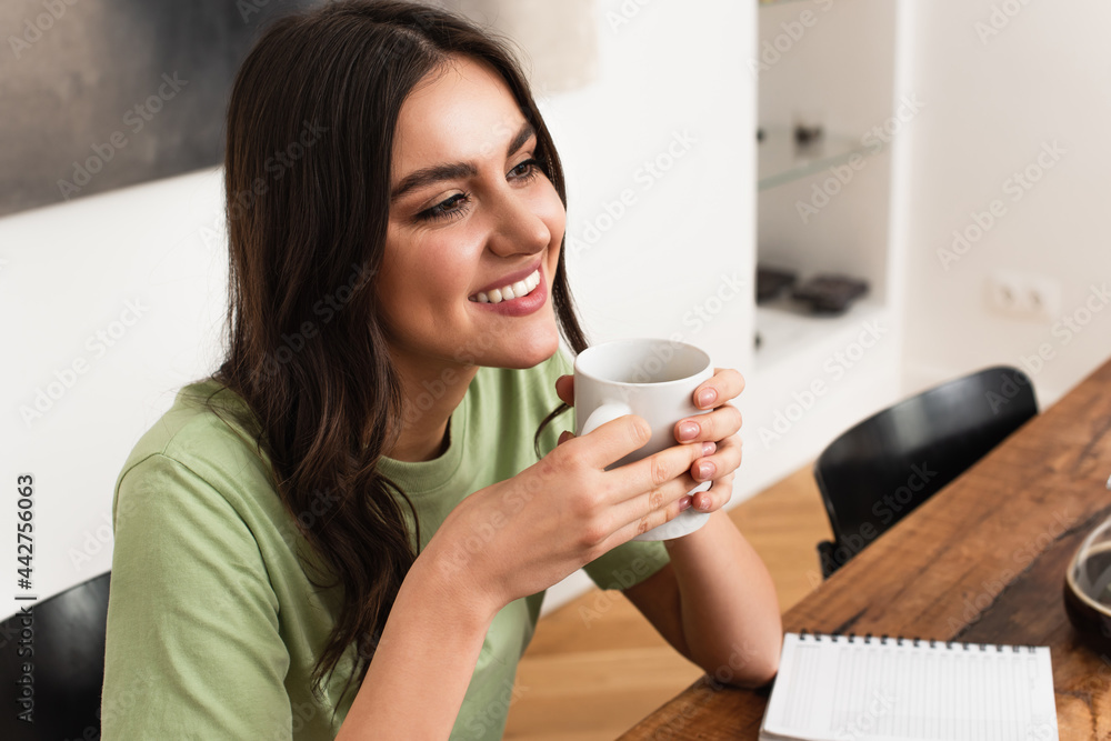 Cheerful woman holding cup of coffee near blurred notebook Cheerful woman holding cup of coffee near blurred notebook