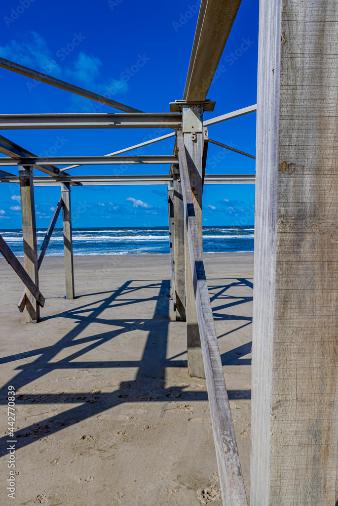 Closeup of wooden posts and beams of an abandoned structure on the beach with the sea with small waves and foam in the background, sunny day in Camperduin, North Holland in the Netherlands