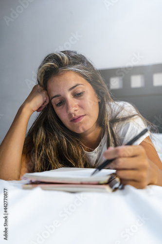 young left-handed hispanic woman lying in bed in her pajamas writing notes in her personal diary