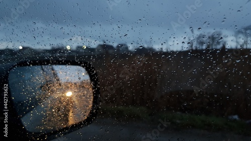 Wet glass texture with raindrops on window. Car side view mirror with defocused reflections and blurred background of autumn night with defocused car lights