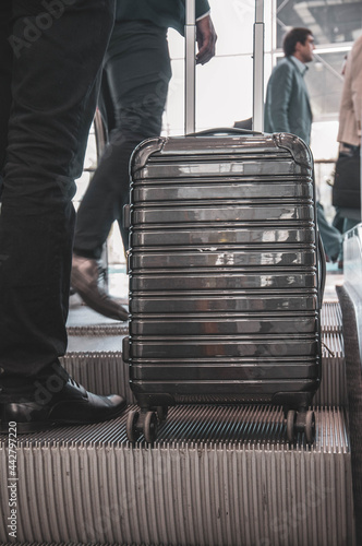 Stylish grey suitcase with wheels on escalator moving stairs near businessman leg in expensive suit and shoes. Business trip concept. Closeup reflective surface of modern hand luggage in airport