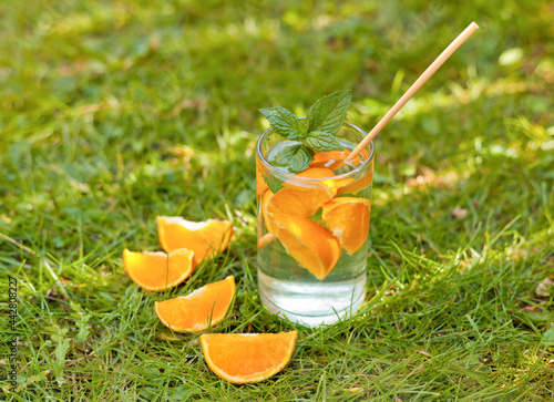 cold lemonade drink with orange and mint in tall glass on green grass and eco straw. glass with oranges and water on the grass