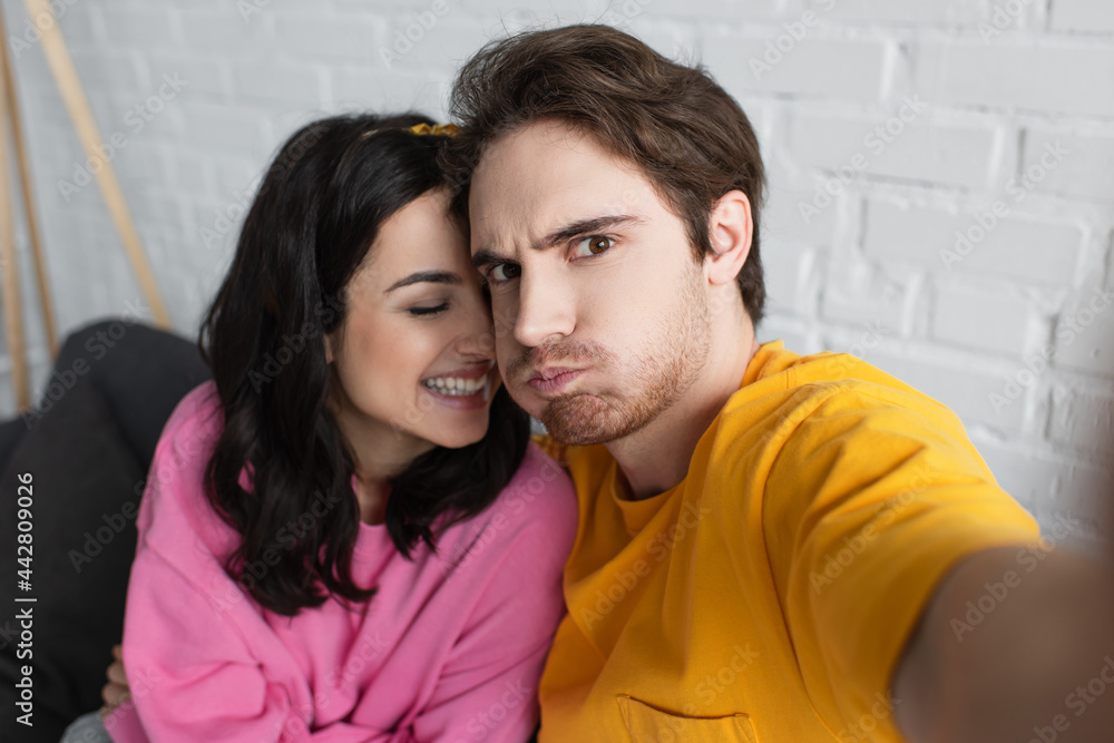 smiling young woman sitting on couch and hugging grimacing boyfriend with outstretched hand in living room smiling young woman sitting on couch and hugging grimacing boyfriend with outstretched hand in living room