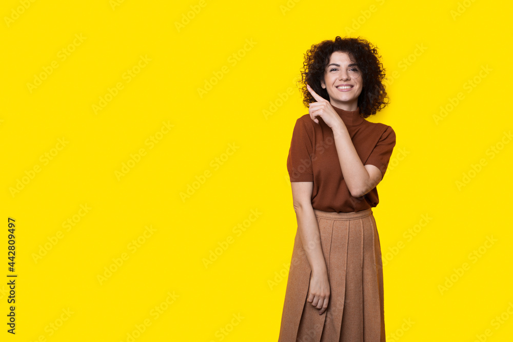 Curly haired lady with nice smile is advertising something on a yellow studio wall by pointing at it Curly haired lady with nice smile is advertising something on a yellow studio wall by pointing at it