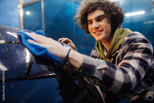 Smiling man in casual clothes cleaning car with rag on blurred foreground