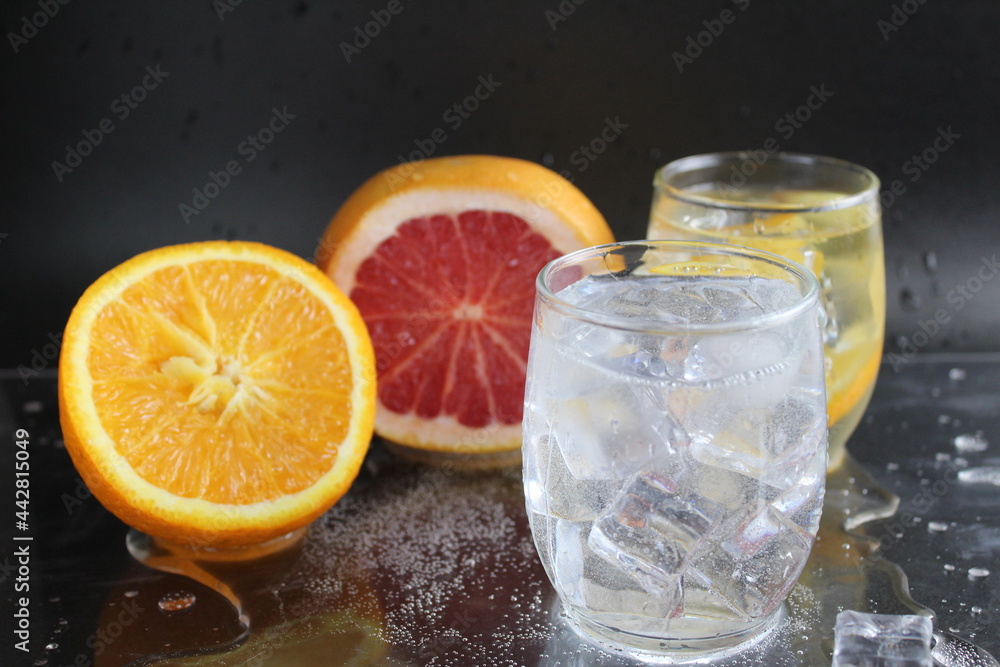 splashes of water over a glass of water and ice. Nearby are fruits orange and grapefruit, ice. On a black background. Refreshing cold ice drink splashes of water over a glass of water and ice. Nearby are fruits orange and grapefruit, ice. On a black background. Refreshing cold ice drink
