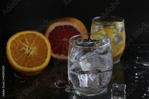 splashes of water over a glass of water and ice. Nearby are fruits orange and grapefruit, ice. On a black background. Refreshing cold ice drink
