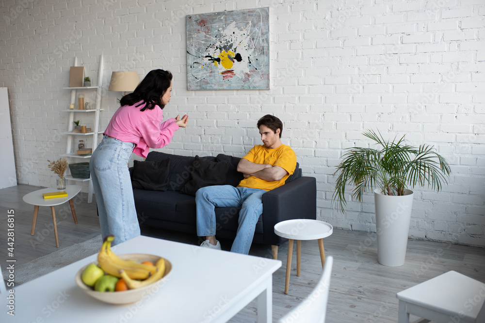 angry young woman gesturing with hands near boyfriend sitting on couch with crossed arms in living room angry young woman gesturing with hands near boyfriend sitting on couch with crossed arms in living room