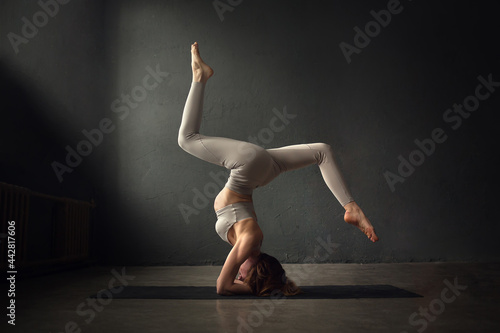 A slim beautifully lighted young  woman is doing shirshasana yoga pose in a dark room. Image with selective focus and toning
